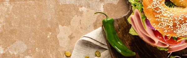 Top view of fresh delicious bagel with sausage on wooden cutting board on aged beige surface with jalapenos, panoramic shot — Stock Photo