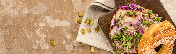 Top view of fresh delicious bagel with meat, red onion and sprouts on wooden cutting board near napkin with pumpkin seeds on aged beige surface, panoramic shot — Stock Photo