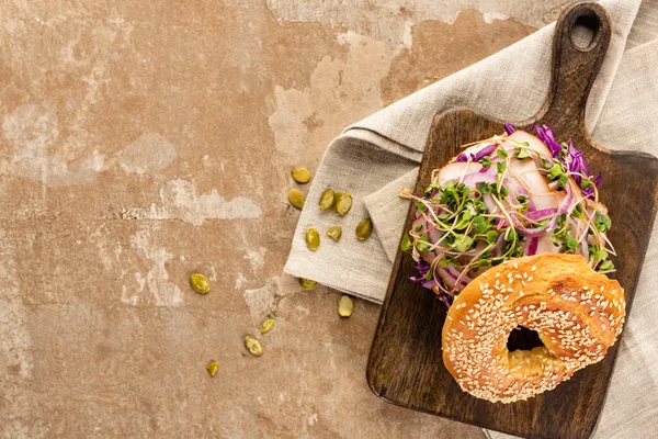 Draufsicht auf frischen leckeren Bagel mit Fleisch, roten Zwiebeln und Rosenkohl auf Holzschneidebrett auf Serviette mit Kürbiskernen auf gealterter beiger Oberfläche — Stockfoto