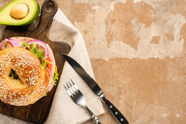 Draufsicht auf frischen leckeren Bagel auf Holzschneidebrett auf gealterter beiger Oberfläche mit Avocado, Besteck und Serviette — Stockfoto