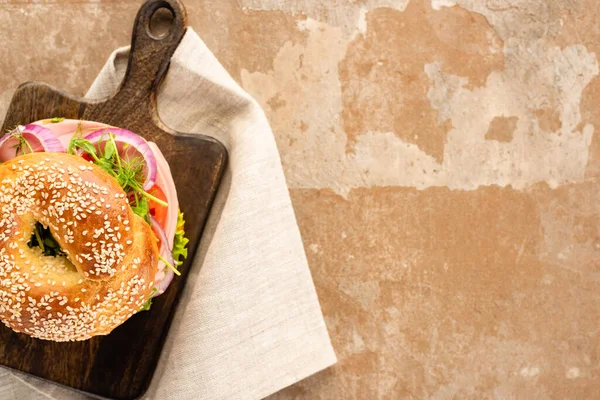 Top view of fresh delicious bagel on wooden cutting board on aged beige surface with napkin — Stock Photo
