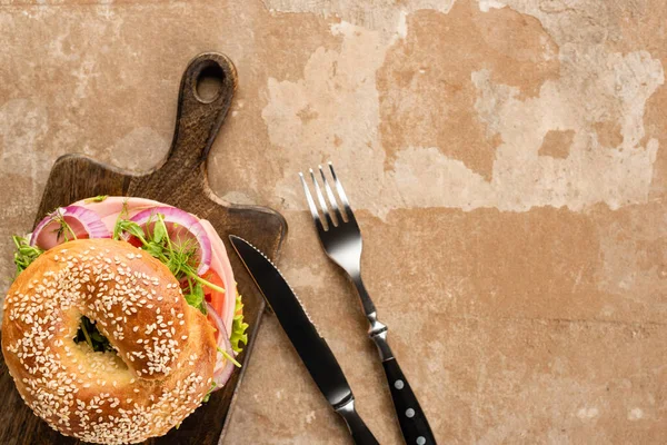 Top view of fresh delicious bagel on wooden cutting board on aged beige surface with cutlery — Stock Photo