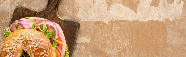 Top view of fresh delicious bagel on wooden cutting board on aged beige surface, panoramic shot — Stock Photo