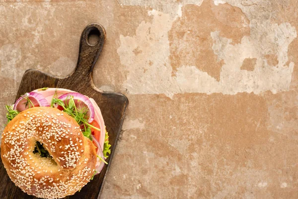 Top view of fresh delicious bagel on wooden cutting board on aged beige surface — Stock Photo