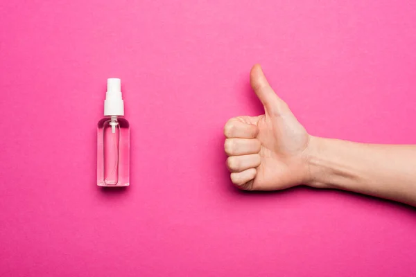 Vista recortada de la mujer que muestra el pulgar hacia arriba cerca de la botella de aerosol con la mano antiséptico en rosa - foto de stock