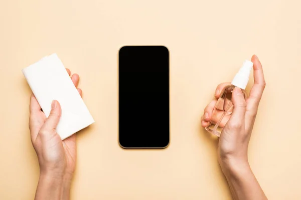Vista parziale di donna che tiene disinfettante e tovaglioli di carta vicino smartphone con schermo bianco su beige — Foto stock