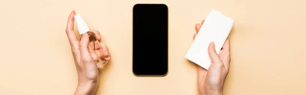 Горизонтальне зображення жіночих рук з дезінфікуючими засобами та паперовими серветками біля смартфона з порожнім екраном на бежевому — стокове фото