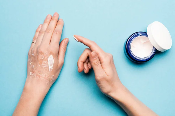 Vista recortada de la mujer aplicando crema de manos sobre la piel deshidratada y exfoliada sobre azul - foto de stock