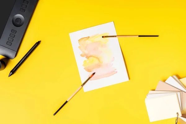 Top view of drawing tablet, painting and paintbrushes near stylus and color palette on yellow — Stock Photo