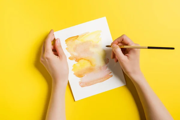 Cropped view of woman holding paintbrush near painting on yellow — Stock Photo