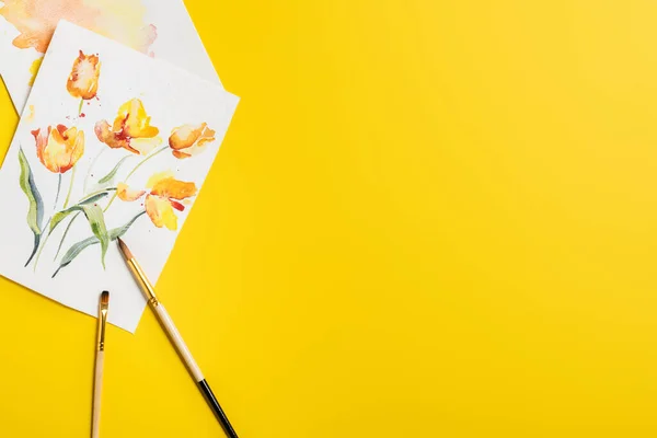 Top view of paintbrushes near creative paintings with drawn flowers isolated on yellow — Stock Photo
