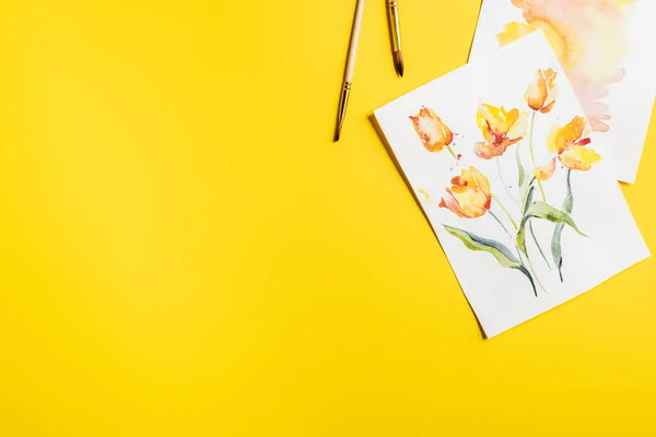 Top view of paintbrushes near paintings with drawn flowers on yellow — Stock Photo