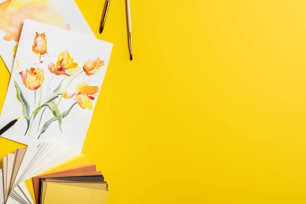 Vista superior de pincéis perto de pinturas com flores desenhadas e paleta de cores em amarelo — Fotografia de Stock