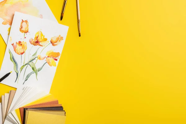 Top view of paintbrushes near paintings with drawn flowers and color palette on yellow — Stock Photo