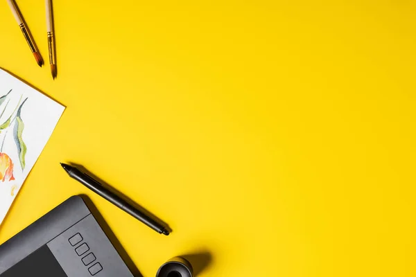 Top view of paintbrushes near painting, drawing tablet, holder and stylus on yellow — Stock Photo