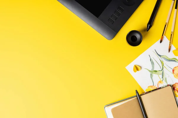 Top view of paintbrushes near painting, drawing tablet, notebook and stylus on yellow — Stock Photo