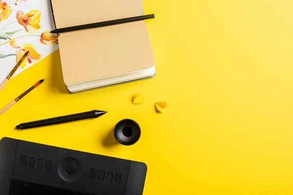 Top view of paintbrushes near painting, drawing tablet, blank notebook and stylus on yellow — Stock Photo