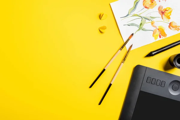 Top view of paintbrushes near painting, paper cut elements, drawing tablet and stylus on yellow — Stock Photo