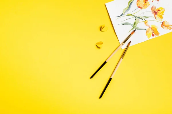 Top view of paintbrushes near drawn flowers on painting and paper cut elements on yellow — Stock Photo