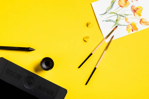 Vue du dessus des pinceaux près des fleurs dessinées sur la peinture, les éléments coupés en papier, la tablette de dessin et le stylet sur le jaune — Photo de stock