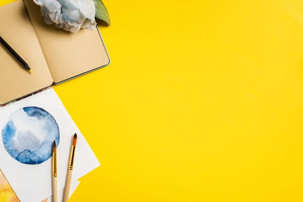 Top view of paintbrushes near paintings, blank notebook and flower on yellow — Stock Photo