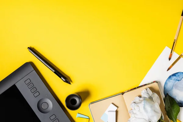 Top view of paintbrushes near painting, drawing tablet, stylus, notebook and flower on yellow — Stock Photo