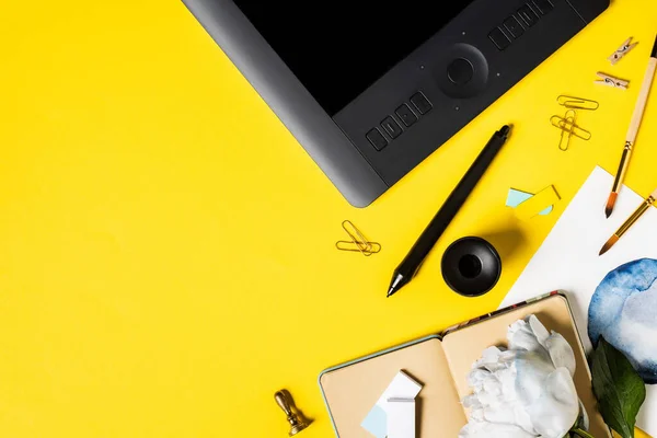 Vue du dessus de la peinture, tablette de dessin avec écran blanc, clips, stylet, cahier et fleur sur jaune — Photo de stock