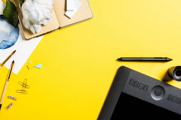 Vista superior de la tableta de dibujo con pantalla en blanco, clips, lápiz óptico, pintura, cuaderno y flor en amarillo - foto de stock