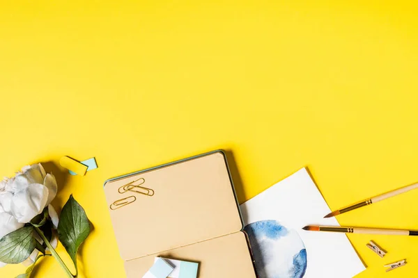 Top view of paintbrushes, clips, painting, blank notebook and flower on yellow — Stock Photo