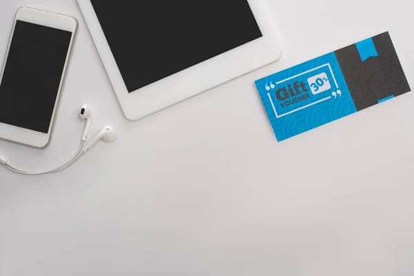 Top view of gift voucher near smartphone, digital tablet and earphones on white surface — Stock Photo