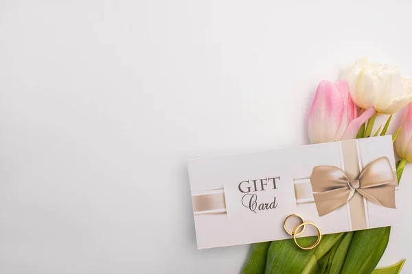 Vista superior de la tarjeta de regalo, anillos de boda y tulipanes sobre fondo blanco - foto de stock
