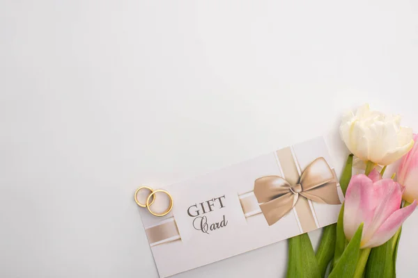 Vista superior de los anillos de boda en la tarjeta de regalo cerca de tulipanes sobre fondo blanco - foto de stock