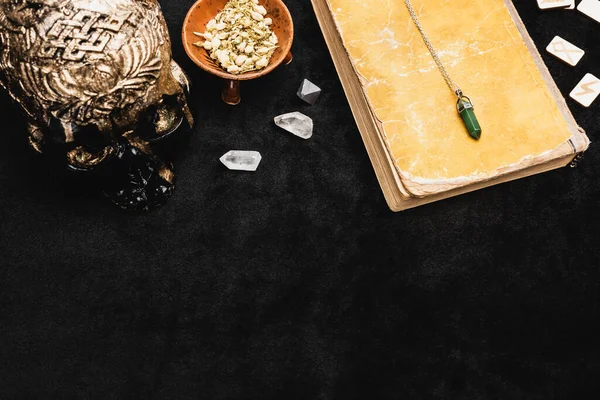 Top view of amulet on aged book near dried herb, occult voodoo skull, runes and crystals on black — Stock Photo