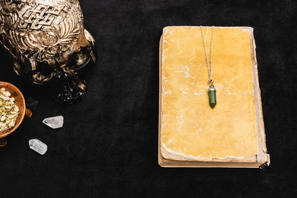 Top view of amulet on aged book near sagebrush, occult voodoo skull, and crystals on black — Stock Photo