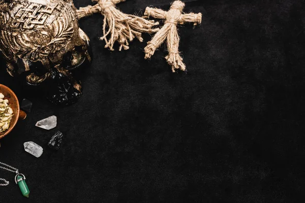 Top view of herb in bowl near voodoo dolls, skull and crystals on black — Stock Photo