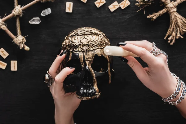 Vue recadrée de la femme tenant une bougie au-dessus du crâne près des runes et des cristaux sur noir — Photo de stock