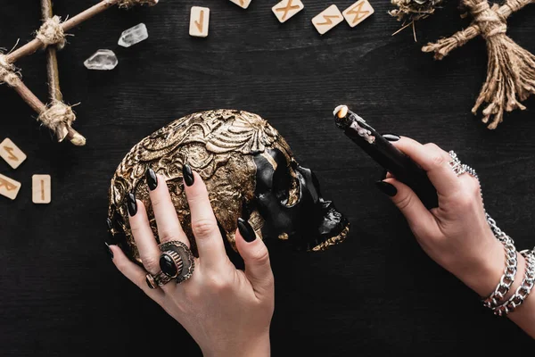 Vue recadrée de la femme tenant une bougie près du crâne, des runes et des cristaux sur noir — Photo de stock