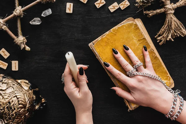 Vista dall'alto della donna che tiene la candela vicino al libro invecchiato, bambola voodoo, rune, cristalli e teschio sul nero — Foto stock