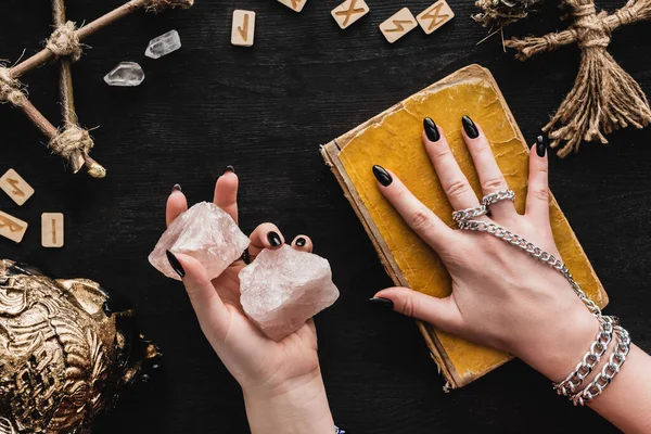 Vue de dessus de sorcière tenant des cristaux et touchant vieux livre de magie noire près de poupée vaudou et runes sur noir — Photo de stock