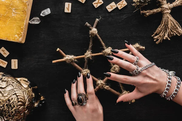 Cropped view of witch touching pentagram near runes, book, crystals, skull and voodoo doll on black — Stock Photo