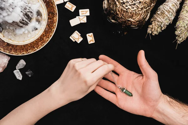 Top view of witch touching amulet on hand of man near runes, wax on candle and skull on black — Stock Photo