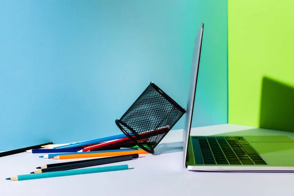 Scattered colored pencils from pencil holder near modern laptop on blue, green and white background — Stock Photo