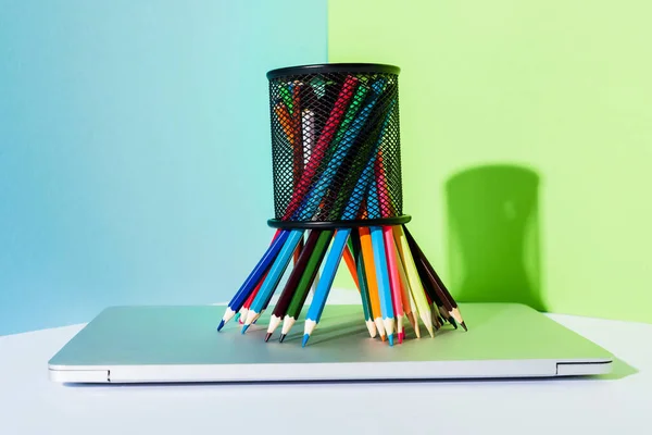 Цветные карандаши в держателе карандаша на современном ноутбуке на синем, зеленом и белом фоне — стоковое фото
