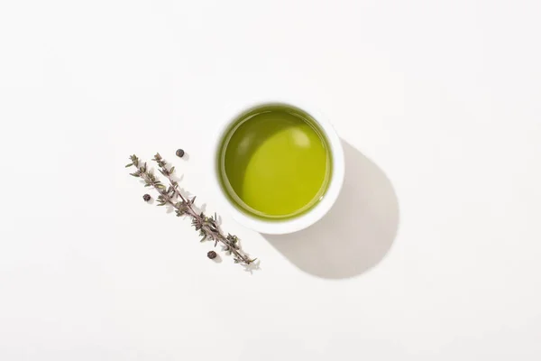 Vue du dessus de l'huile d'olive dans un bol près de l'herbe et du poivre noir sur fond blanc — Photo de stock