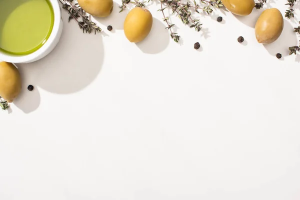 Вид зверху на оливкову олію в мисці біля зелених оливок, трави та чорного перцю на білому тлі — стокове фото