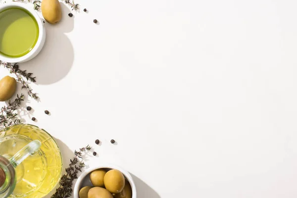 Vista dall'alto dell'olio d'oliva in ciotola e bottiglia vicino a olive verdi, erbe aromatiche e pepe nero su sfondo bianco — Foto stock