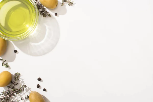 Vista dall'alto dell'olio in una ciotola di vetro vicino a olive verdi, erbe aromatiche e pepe nero su sfondo bianco — Foto stock