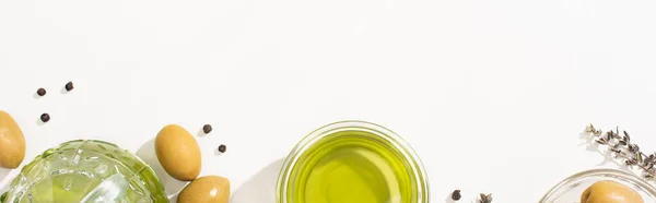 Вид зверху на оливкову олію в мисці і пляшці біля зелених оливок, трава і чорний перець на білому тлі, панорамна орієнтація — стокове фото