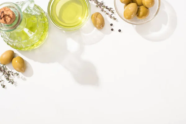 Vista superior del aceite de oliva en tazón y botella cerca de aceitunas verdes, hierba y pimienta negra sobre fondo blanco - foto de stock