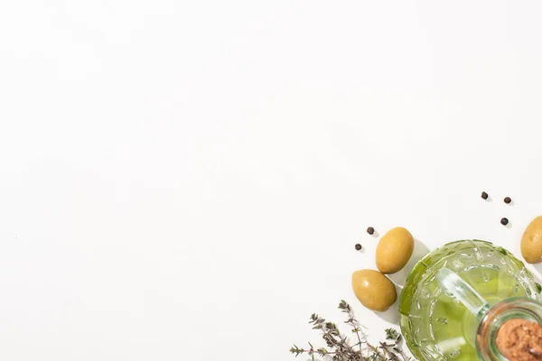 Ansicht von Olivenöl in der Flasche in der Nähe von grünen Oliven, Kräutern und schwarzem Pfeffer auf weißem Hintergrund — Stockfoto